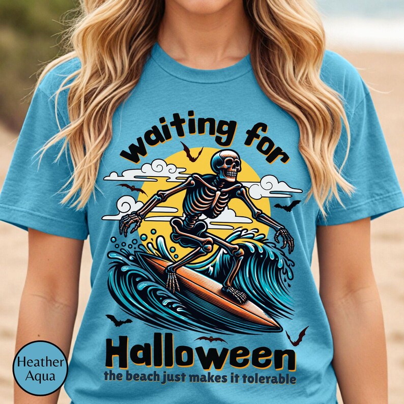 May include: Heather aqua t-shirt with a Halloween theme. The shirt features a skeleton surfing on a wave with the words "waiting for Halloween" and "the beach just makes it tolerable".