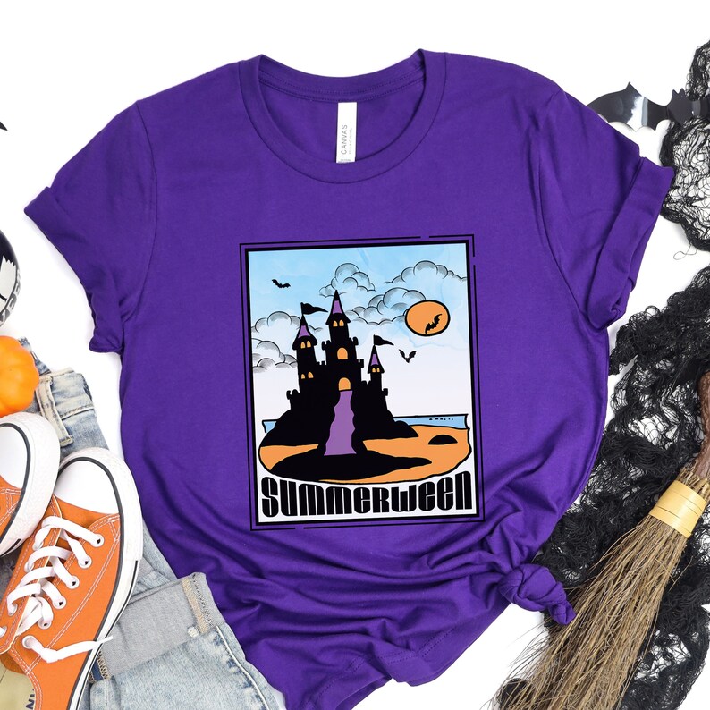 Pu&ograve; includere: Maglietta viola con una grafica di un castello nero su una spiaggia con il testo "Summerween".