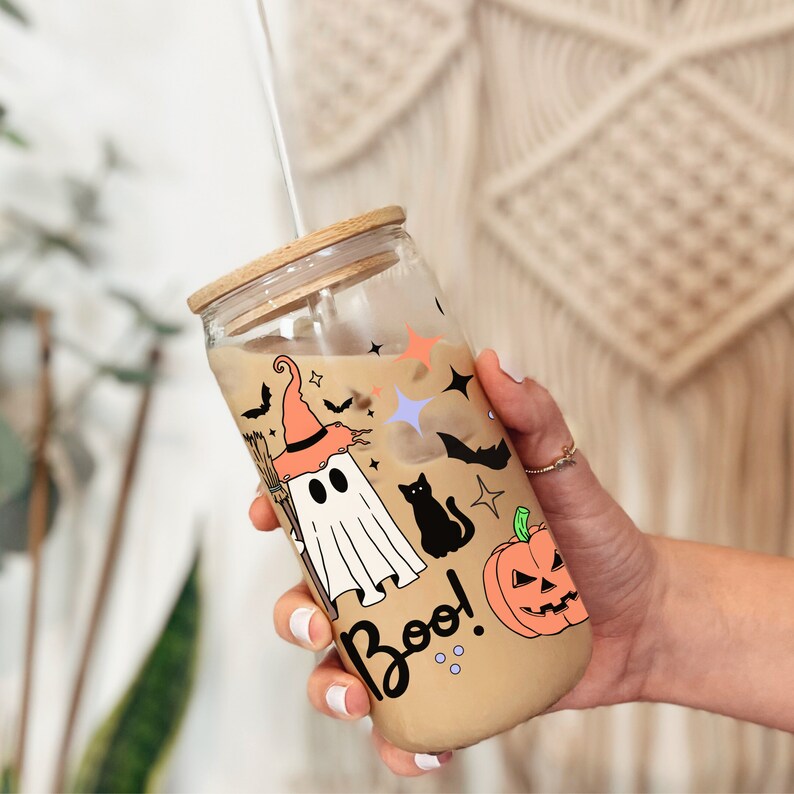 May include: A clear glass reusable tumbler with a bamboo lid and a straw. The tumbler has a Halloween theme with a ghost, a black cat, a pumpkin, and the word "Boo!" printed on it.