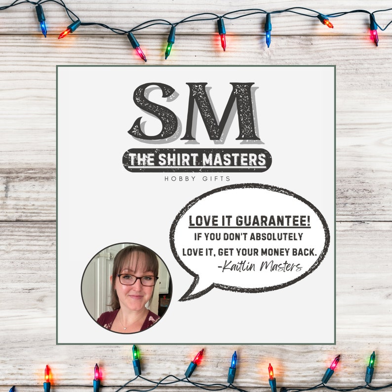 May include: A white sign with black text that reads "SM THE SHIRT MASTERS HOBBY GIFTS". Below the text is a speech bubble with the text "LOVE IT GUARANTEE! IF YOU DON'T ABSOLUTELY LOVE IT, GET YOUR MONEY BACK. -Kaitlin Masters". A woman with glasses is pictured in the background.