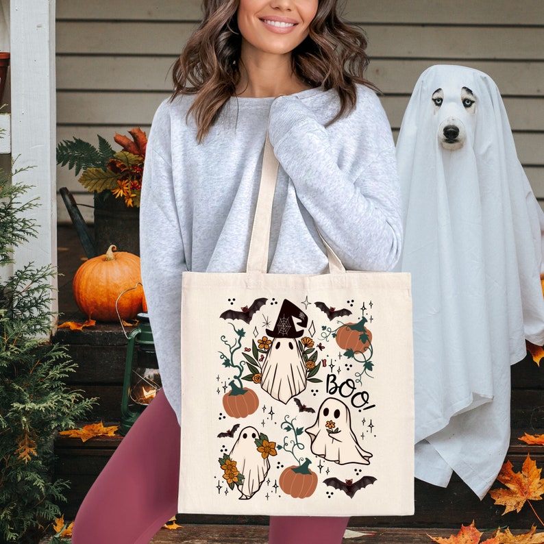 K&ouml;nnte beinhalten: Beige Halloween-Tragetasche mit Geisterillustrationen, K&uuml;rbissen, Flederm&auml;usen und dem Wort "Boo!". Das Design beinhaltet einen Geist mit Hexenhut. Die Tasche wird von einer Person gehalten.