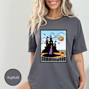 Pu&ograve; includere: Una maglietta grigia con una grafica di un castello nero su una spiaggia con il testo "Summerween" in arancione e nero.
