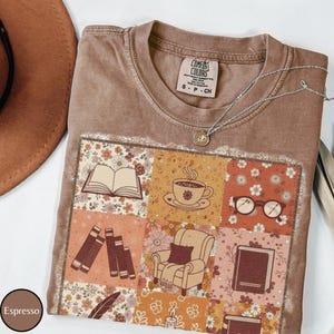 May include: Espresso-colored t-shirt with a patchwork design featuring books, a teacup, glasses, and a cozy armchair. A silver necklace with a small pendant rests on the shirt. The shirt has a Comfort Colors label.