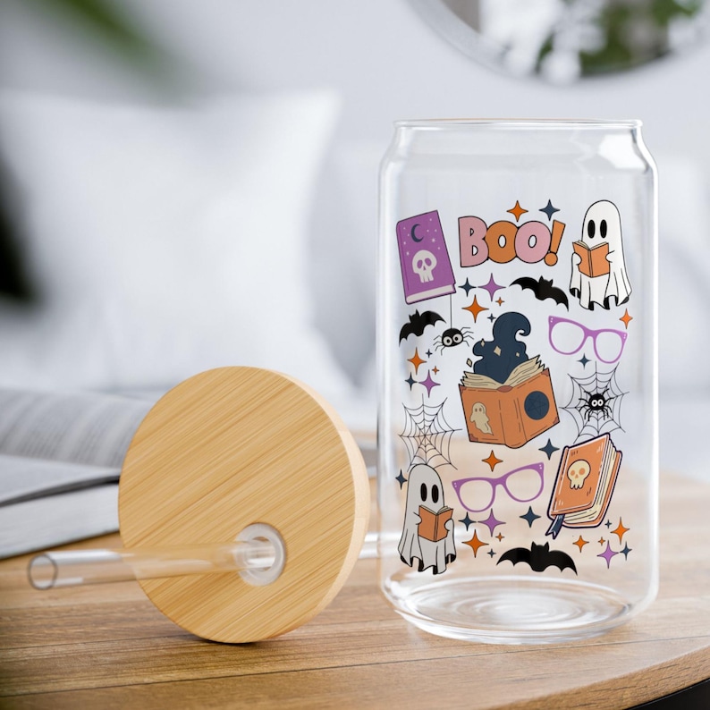Puede incluir: Un vaso de cristal transparente con forma de lata, tapa de bamb&uacute; y pajita de cristal. El vaso presenta un estampado colorido de fantasmas, libros, ara&ntilde;as, telara&ntilde;as, murci&eacute;lagos y gafas, con el texto "BOO!" en letras rosas y peque&ntilde;as estrellas.