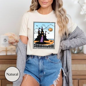 Pu&ograve; includere: Una maglietta bianca con una grafica di un castello nero su una spiaggia con il testo "Summerween" in un font retr&ograve;.
