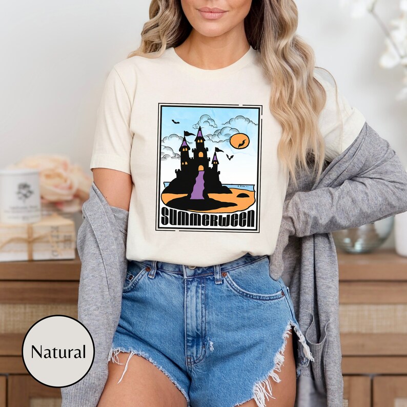 Pu&ograve; includere: Una maglietta bianca con una grafica di un castello nero su una spiaggia con il testo "Summerween" in un font retr&ograve;.