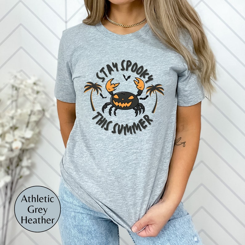以下が含まれることがあります： グレーヘザーのTシャツに、黒とオレンジのグラフィックで、ジャック・オー・ランタンの顔をしたカニが描かれています。カニの周りに「Stay Spooky This Summer」という文字が印刷されています。