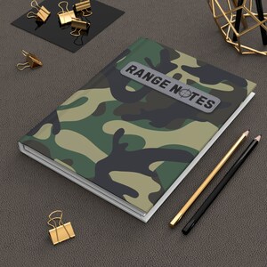 May include: A camouflage notebook with a white border and a gray label that says "RANGE NOTES" with a target symbol. The notebook is on a brown leather surface with two pencils and gold paper clips.
