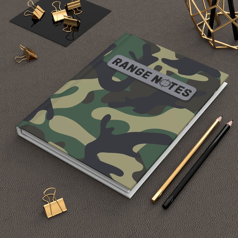 May include: A camouflage notebook with a white border and a gray label that says "RANGE NOTES" with a target symbol. The notebook is on a brown leather surface with two pencils and gold paper clips.