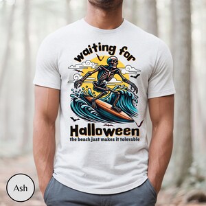 May include: Ash-colored t-shirt featuring a surfing skeleton graphic. The design includes the words "waiting for Halloween" above the skeleton and "the beach just makes it tolerable" below. The skeleton is riding a surfboard on a wave.