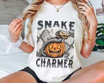 Könnte beinhalten: Cremefarbenes T-Shirt mit einem Halloween-Motiv. Das Design zeigt eine schwarze Schlange, die sich um einen Kürbis mit leuchtenden Augen und einem bedrohlichen Grinsen windet. Die Worte "SNAKE CHARMER" sind oberhalb und unterhalb des Bildes gedruckt.