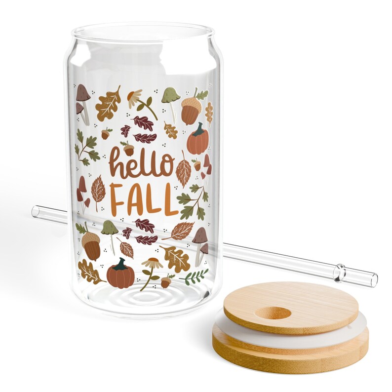 May include: A clear glass can with a straw and a lid. The can has a design of fall leaves, pumpkins, acorns, and mushrooms. The text "hello fall" is printed on the can.