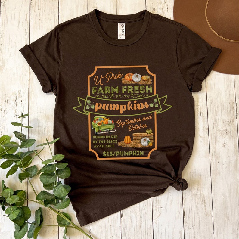 May include: A brown t-shirt with a graphic of a sign that says "U Pick Farm Fresh Pumpkins". The sign is decorated with pumpkins, hay bales, and a truck. The text on the sign also says "September and October" and "$15/Pumpkin".