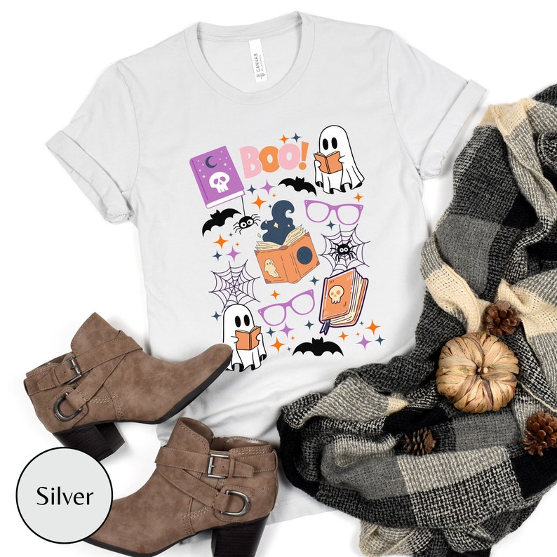 May include: White t-shirt with a Halloween-themed design featuring ghosts, bats, spiders, books, and the word "BOO!" in pink letters.