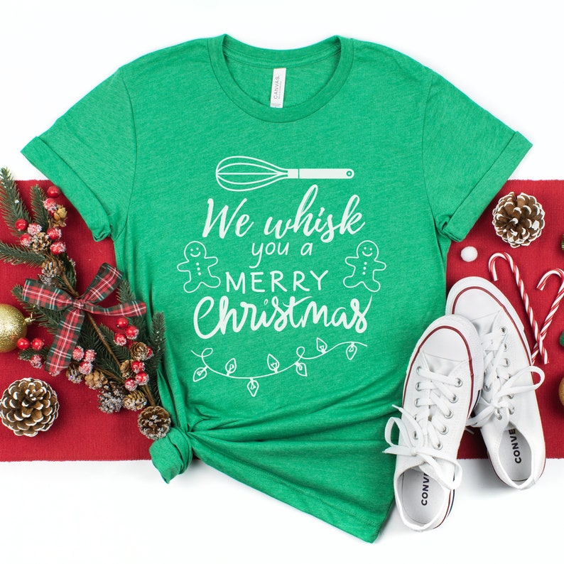 K&ouml;nnte beinhalten: Gr&uuml;nes T-Shirt mit einer wei&szlig;en Grafik, auf der "We whisk you a Merry Christmas" steht. Die Grafik enth&auml;lt einen Schneebesen, zwei Lebkuchenm&auml;nner und eine Lichterkette.