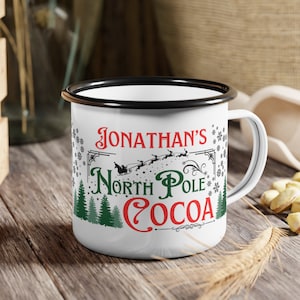 May include: White enamel mug with a black rim. The mug has a red and green design with the text "Jonathan's North Pole Cocoa" and snowflakes.