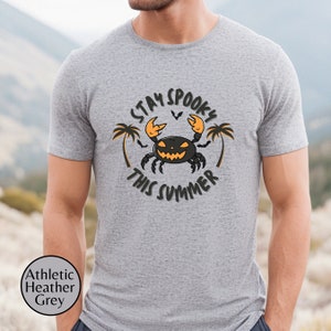 以下が含まれることがあります： グレーのTシャツに、黒とオレンジの蟹のデザインが施されています。蟹はジャック・オー・ランタンの顔をしていて、ヤシの木に囲まれています。蟹の周りに「Stay Spooky This Summer」という文字が書かれています。