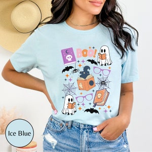 May include: A light blue t-shirt with a Halloween-themed design featuring ghosts, bats, spiders, books, glasses, and the word "Boo!" printed on it.