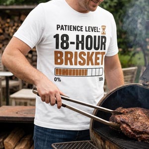 Pode incluir: Camiseta branca com o texto "PATIENCE LEVEL: 18-HOUR BRISKET" em preto e castanho. Um gráfico de barra de progresso mostra de 0% a 18h. A camiseta está sendo usada enquanto grelha um grande pedaço de carne com pinças.