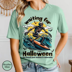 May include: Heather mint green t-shirt with a Halloween-themed graphic. The design features a skeleton surfing a wave with a yellow sun in the background. Text reads "waiting for Halloween" and "the beach just makes it tolerable."