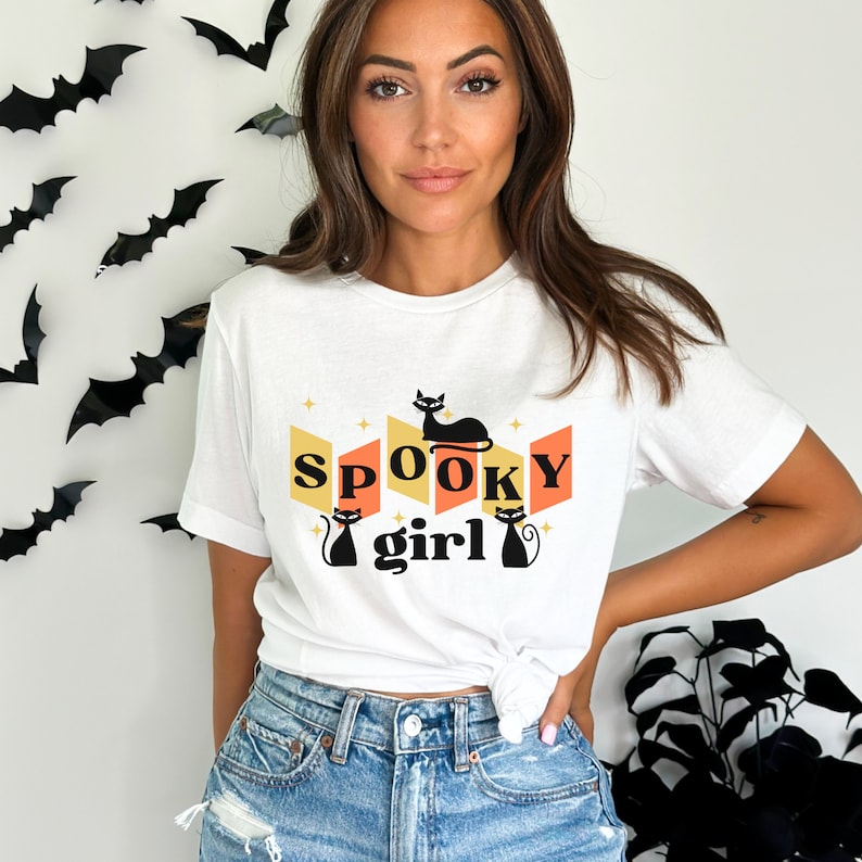 May include: White t-shirt with a retro design featuring the text "SPOOKY girl" and black cat illustrations. The design is in orange, yellow, and white.