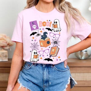 May include: Pink t-shirt with a Halloween-themed design featuring ghosts, bats, spiders, books, and the text "BOO!"