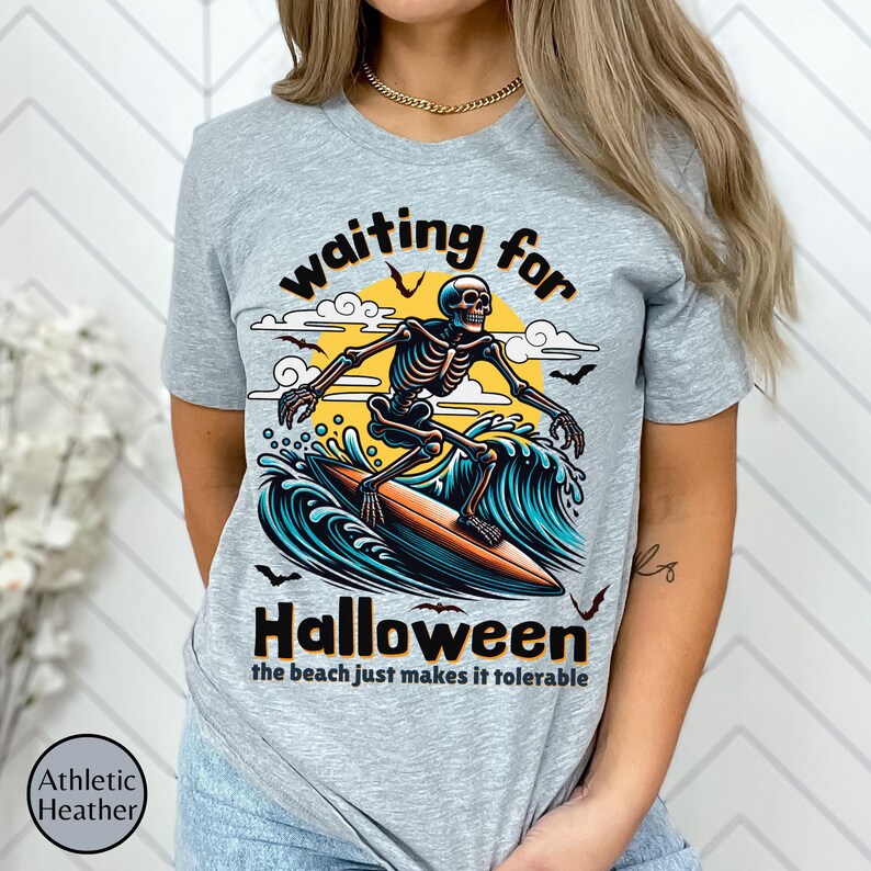 May include: Athletic heather gray t-shirt with a Halloween-themed graphic. The design features a skeleton surfing a wave with the text "waiting for Halloween" and "the beach just makes it tolerable."
