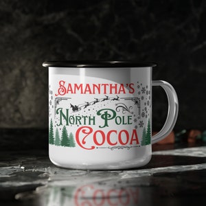 May include: White enamel mug with a black rim and handle. It has a Christmas-themed design with green trees, snowflakes, and the text "Samantha's North Pole Cocoa" in red and black.