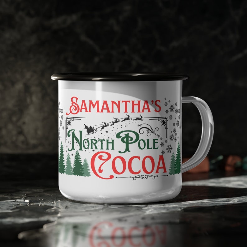 May include: White enamel mug with a black rim and handle. It has a Christmas-themed design with green trees, snowflakes, and the text "Samantha's North Pole Cocoa" in red and black.