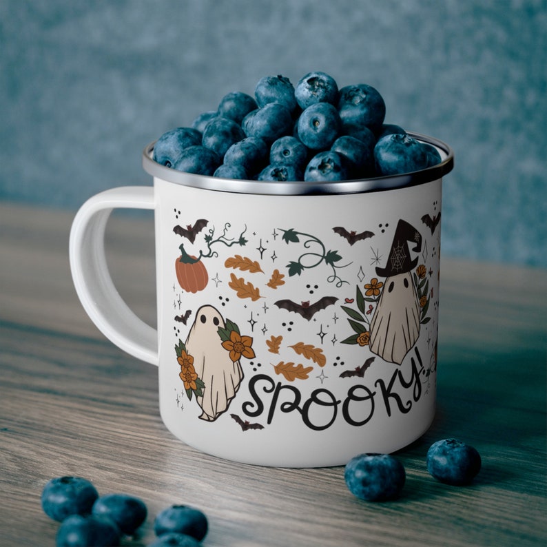May include: White enamel mug with a spooky Halloween design featuring ghosts, pumpkins, bats, and the word "Spooky!"  The mug is filled with blueberries.