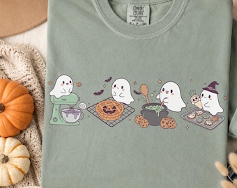 May include: A sage green t-shirt featuring a graphic of four white ghosts baking. The ghosts are shown using a stand mixer, cooling a pumpkin pie, stirring a green potion in a cauldron, and holding a tray of ghost-shaped cookies.