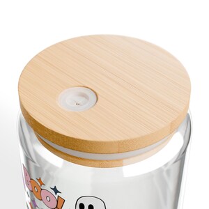 Puede incluir: Un vaso de vidrio transparente con una tapa de bamb&uacute; de color claro que incluye un orificio circular de silicona blanca para pajita. La superficie del vidrio muestra un gr&aacute;fico con la palabra BOO en letras coloridas y un fantasma.