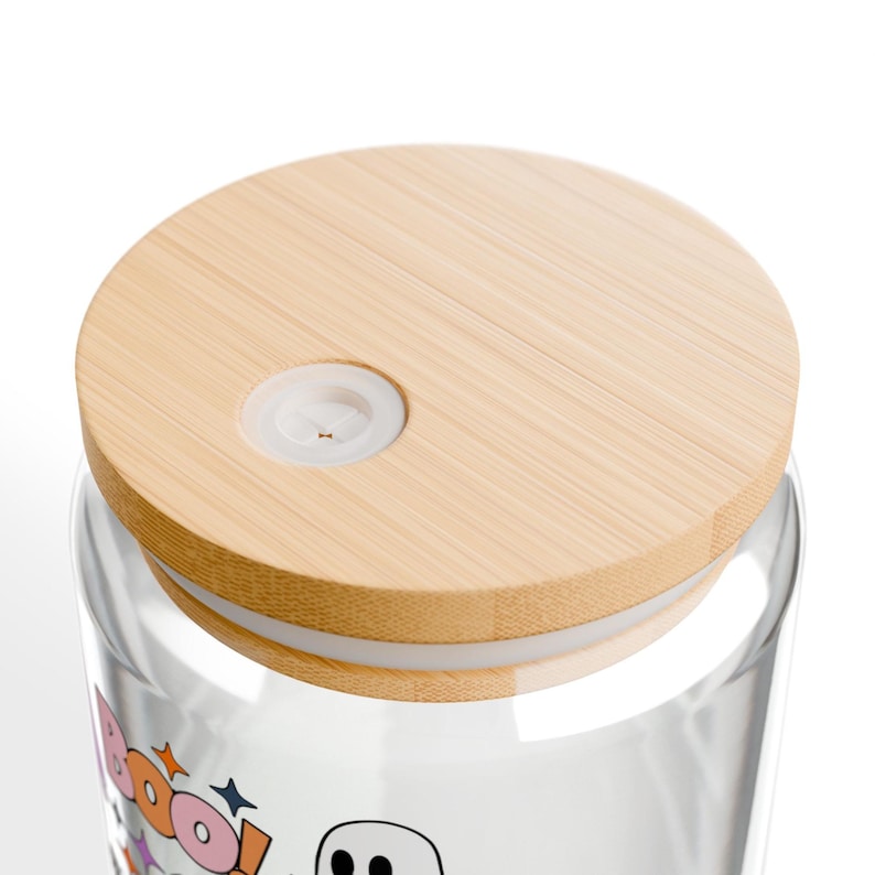 Puede incluir: Un vaso de vidrio transparente con una tapa de bamb&uacute; de color claro que incluye un orificio circular de silicona blanca para pajita. La superficie del vidrio muestra un gr&aacute;fico con la palabra BOO en letras coloridas y un fantasma.