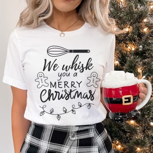 K&ouml;nnte beinhalten: Wei&szlig;es T-Shirt mit einem schwarzen Schneebesen, zwei Lebkuchenm&auml;nnern und dem Text "We whisk you a merry Christmas".