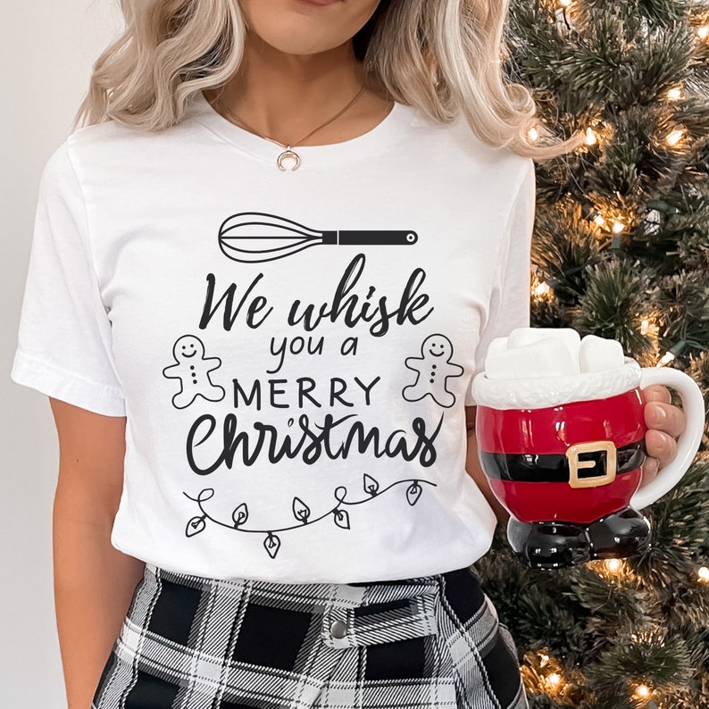 K&ouml;nnte beinhalten: Wei&szlig;es T-Shirt mit einem schwarzen Schneebesen, zwei Lebkuchenm&auml;nnern und dem Text "We whisk you a merry Christmas".