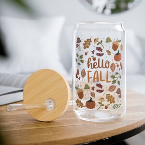 May include: A glass can with a bamboo lid and a straw. The can has a fall-themed design with leaves, acorns, mushrooms, and pumpkins. The text "hello fall" is printed on the can.