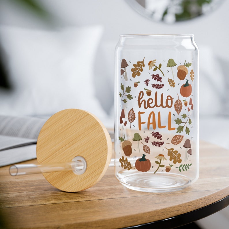 May include: A glass can with a bamboo lid and a straw. The can has a fall-themed design with leaves, acorns, mushrooms, and pumpkins. The text "hello fall" is printed on the can.