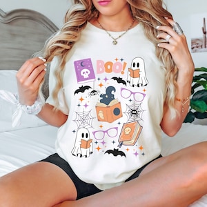 May include: White t-shirt with a Halloween-themed design featuring ghosts, bats, spiders, books, and the word "BOO!" in pink letters.