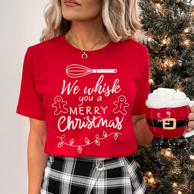 K&ouml;nnte beinhalten: Rotes T-Shirt mit einer wei&szlig;en Grafik, auf der "We whisk you a merry Christmas" mit einem Schneebesen, Lebkuchenm&auml;nnern und Weihnachtslichtern steht.