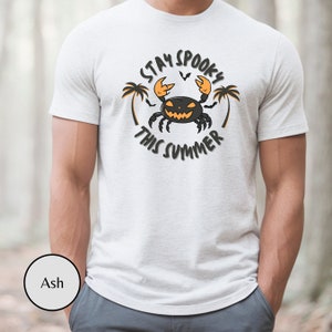 以下が含まれることがあります： 黒いオレンジ色のカニがジャック・オー・ランタンの顔をしている白いTシャツ。カニの周りに円形で「Stay Spooky This Summer」という文字が書かれている。