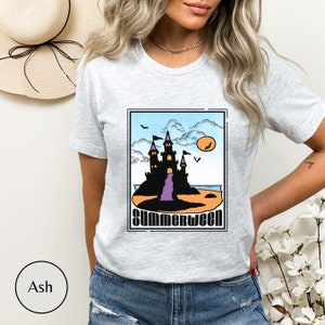 Pu&ograve; includere: Una maglietta grigia chiaro con una stampa grafica di un castello nero su una spiaggia con un ponte viola, un sole arancione e pipistrelli. Il testo "Summerween" &egrave; stampato sotto il castello.