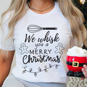 K&ouml;nnte beinhalten: Wei&szlig;es T-Shirt mit einer schwarzen Grafik, auf der "We whisk you a Merry Christmas" mit einem Schneebesen, Lebkuchenm&auml;nnern und Weihnachtslichtern steht.