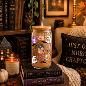 Puede incluir: Un vaso transparente con tapa de bamb&uacute; y pajita de cristal, lleno de una bebida de color marr&oacute;n claro. El exterior presenta calcoman&iacute;as coloridas con tem&aacute;tica de Halloween, incluyendo fantasmas, libros, ara&ntilde;as, telara&ntilde;as, murci&eacute;lagos y BOO.