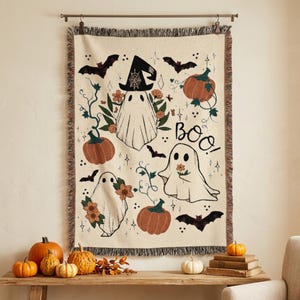 Puede incluir: Una manta tejida de color crema con un patr&oacute;n tem&aacute;tico de Halloween que incluye tres fantasmas blancos, calabazas naranjas, murci&eacute;lagos negros y detalles florales. El texto "BOO!" est&aacute; impreso en negro. La manta tiene un borde con flecos.