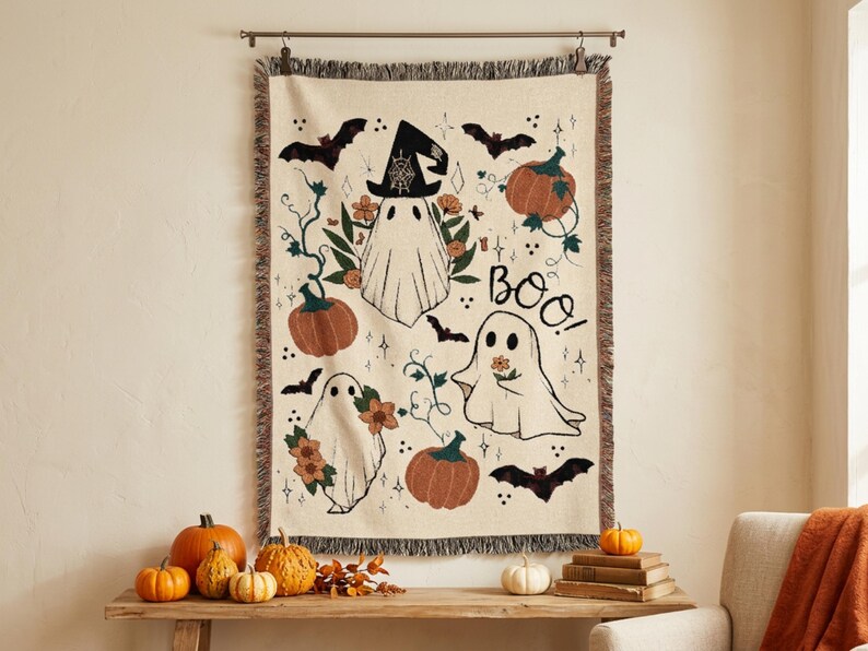 Puede incluir: Una manta tejida de color crema con un patr&oacute;n tem&aacute;tico de Halloween que incluye tres fantasmas blancos, calabazas naranjas, murci&eacute;lagos negros y detalles florales. El texto "BOO!" est&aacute; impreso en negro. La manta tiene un borde con flecos.