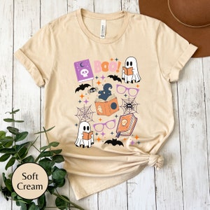 May include: A soft cream colored t-shirt with a Halloween themed graphic featuring ghosts, bats, spiders, and books. The text "BOO!" is printed on the shirt.