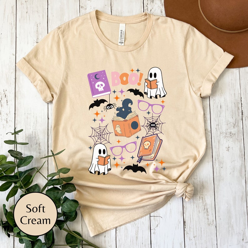 May include: A soft cream colored t-shirt with a Halloween themed graphic featuring ghosts, bats, spiders, and books. The text "BOO!" is printed on the shirt.