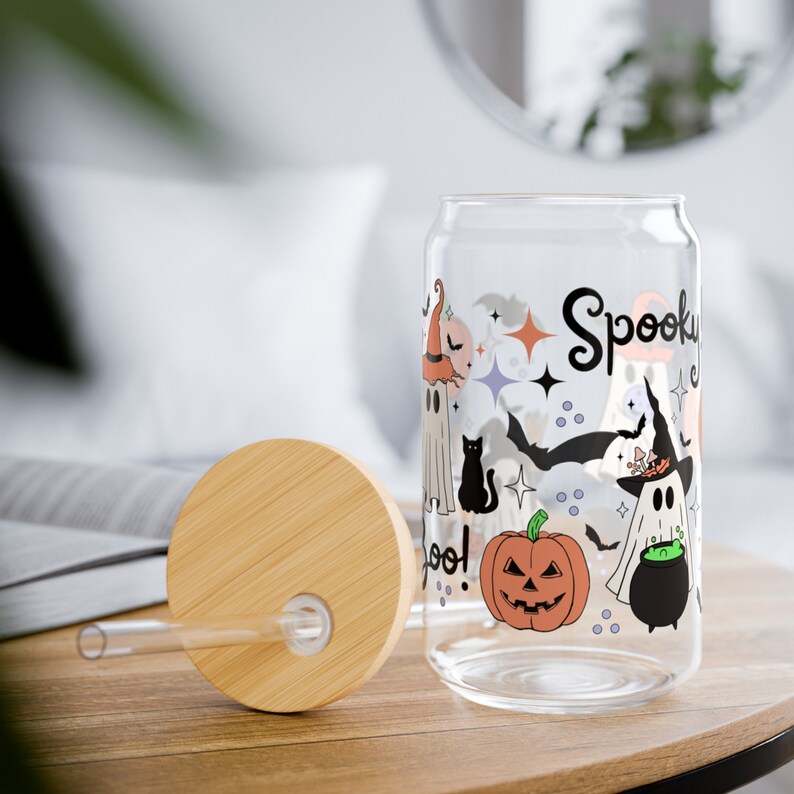 May include: A clear glass can with a spooky Halloween design. The can features ghosts, bats, a black cat, a pumpkin, and a cauldron. The text "Spooky" is written in a whimsical font on the can.