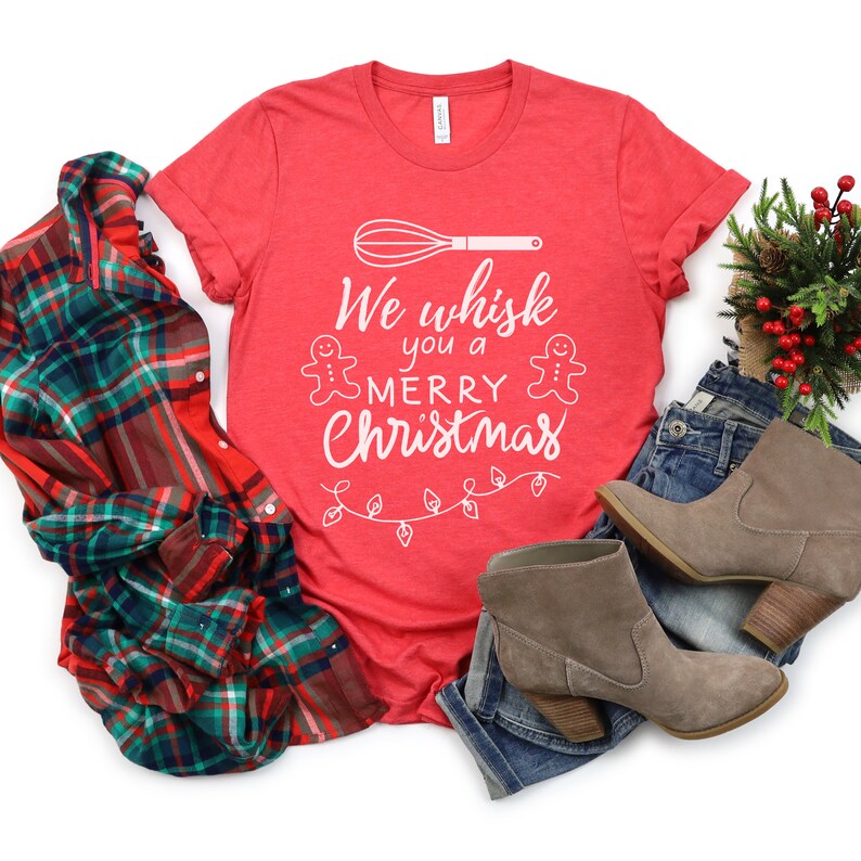 K&ouml;nnte beinhalten: Rotes T-Shirt mit einer wei&szlig;en Grafik, auf der "We whisk you a Merry Christmas" mit einem Schneebesen, Lebkuchenm&auml;nnern und Weihnachtslichtern steht.