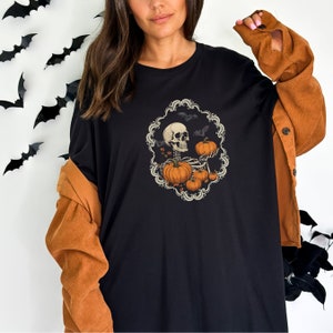 Op de afbeelding: Zwart T-shirt met een afbeelding van een skelet dat pompoenen vasthoudt. De afbeelding is omgeven door een decoratief frame met vleermuizen en bladeren.